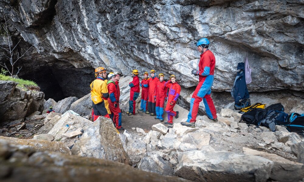 Eine Gruppe von Personen mit Helmen und Schutzanzügen steht in der Nähe des Eingangs einer Felsenhöhle und hört einer Person zu, die spricht. In der Nähe ist Ausrüstung auf dem Boden zu sehen.