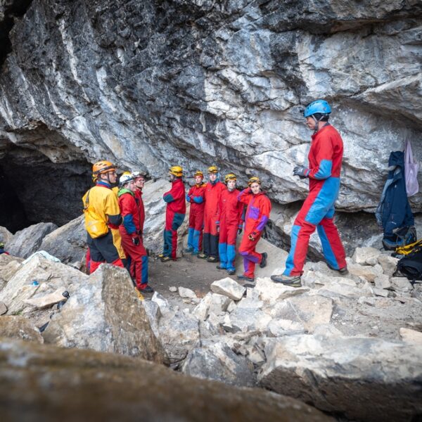 Eine Gruppe von Personen mit Helmen und Schutzanzügen steht in der Nähe des Eingangs einer Felsenhöhle und hört einer Person zu, die spricht. In der Nähe ist Ausrüstung auf dem Boden zu sehen.