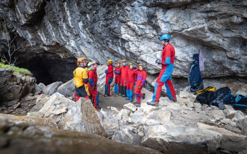 Eine Gruppe von Personen mit Helmen und Schutzanzügen steht in der Nähe des Eingangs einer Felsenhöhle und hört einer Person zu, die spricht. In der Nähe ist Ausrüstung auf dem Boden zu sehen.