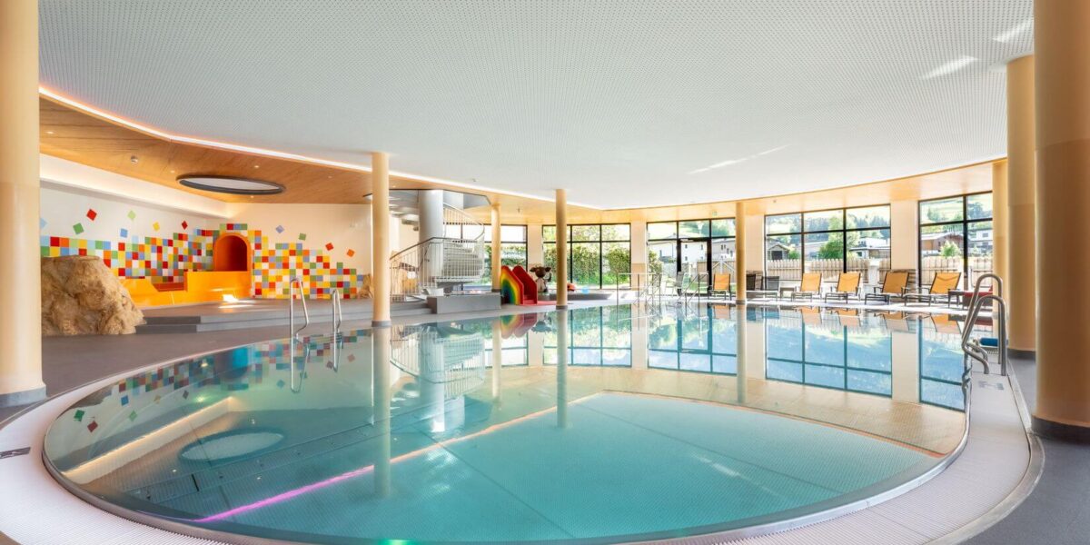 Modernes Hallenbad mit rundem Becken, Liegestühlen an großen Fenstern und einem farbenfrohen Spielbereich für Kinder im Hintergrund.