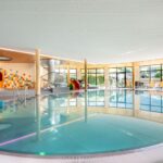 Modernes Hallenbad mit rundem Becken, Liegestühlen an großen Fenstern und einem farbenfrohen Spielbereich für Kinder im Hintergrund.