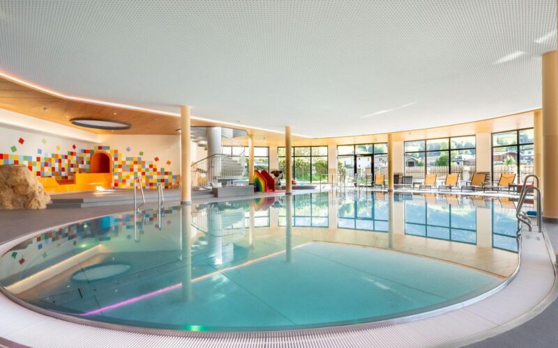 Modernes Hallenbad mit rundem Becken, Liegestühlen an großen Fenstern und einem farbenfrohen Spielbereich für Kinder im Hintergrund.