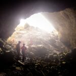 Mehrere Personen mit Helmen und roter Kleidung stehen in einer großen, felsigen Höhle, in die Licht von oben einfällt.