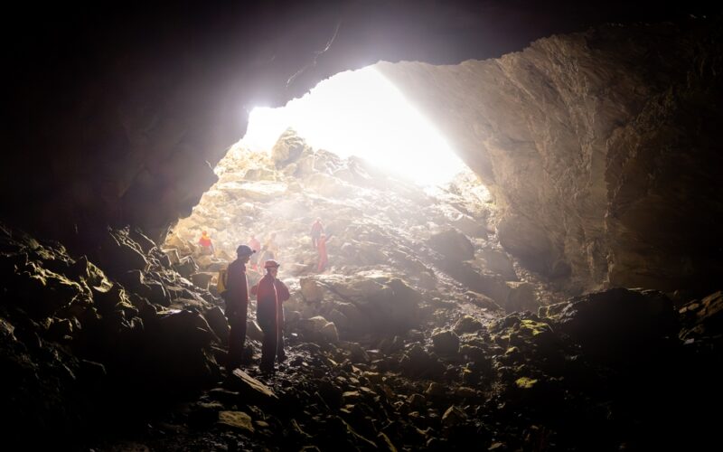 Mehrere Personen mit Helmen und roter Kleidung stehen in einer großen, felsigen Höhle, in die Licht von oben einfällt.