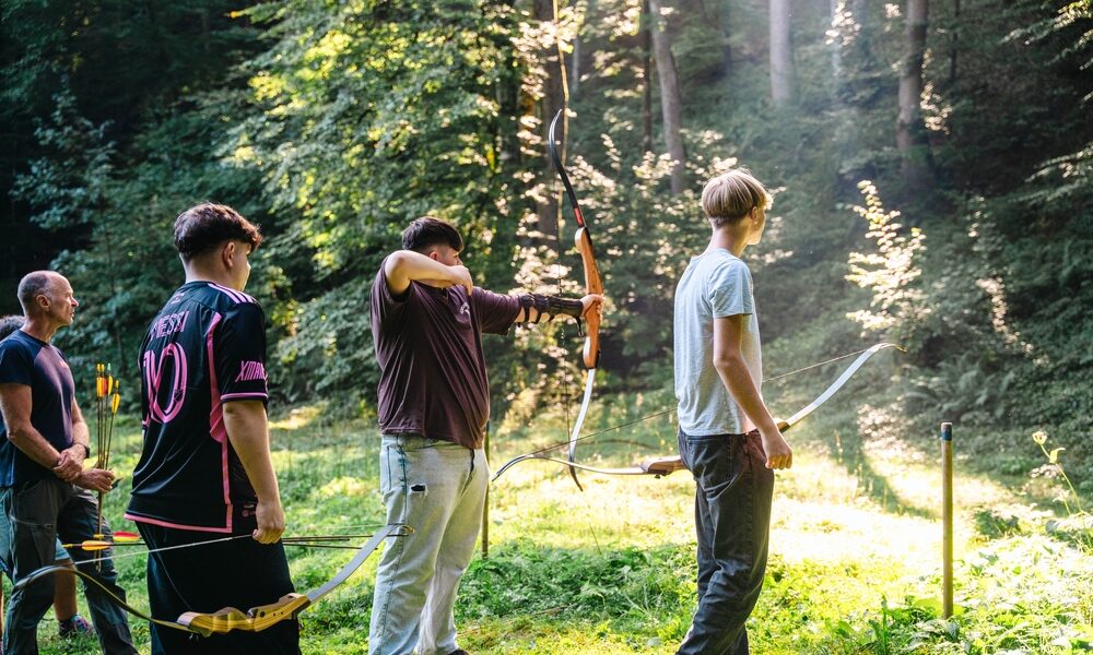 Vier Personen stehen auf einer sonnenbeschienenen Waldlichtung, drei von ihnen halten Pfeil und Bogen, eine Person zielt auf ein Ziel außerhalb des Bildes.