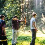 Vier Personen stehen auf einer sonnenbeschienenen Waldlichtung, drei von ihnen halten Pfeil und Bogen, eine Person zielt auf ein Ziel außerhalb des Bildes.