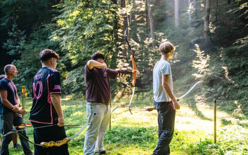 Vier Personen stehen auf einer sonnenbeschienenen Waldlichtung, drei von ihnen halten Pfeil und Bogen, eine Person zielt auf ein Ziel außerhalb des Bildes.