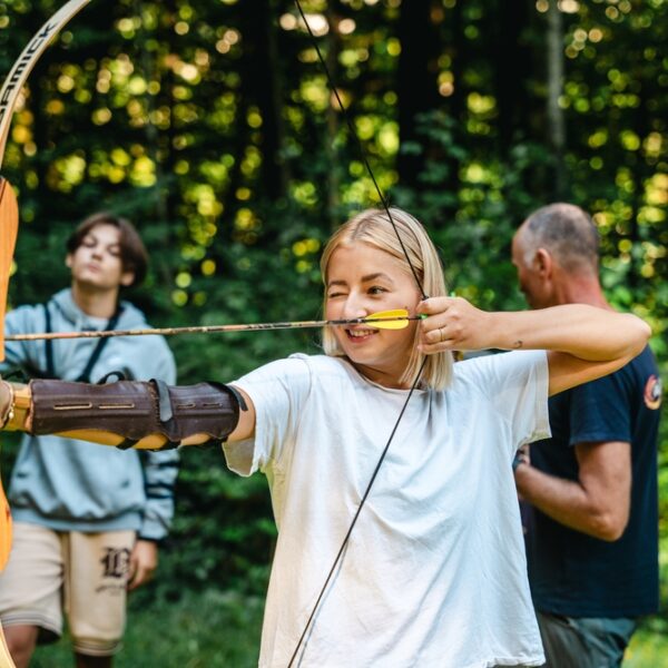 Ein junger Mensch zielt mit Pfeil und Bogen im Freien, während andere in der Nähe sind. Bäume und Grünpflanzen füllen den Hintergrund.