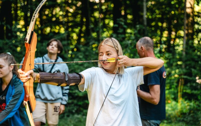 Ein junger Mensch zielt mit Pfeil und Bogen im Freien, während andere in der Nähe sind. Bäume und Grünpflanzen füllen den Hintergrund.