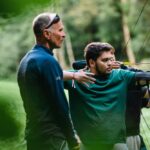 Ein Mann unterrichtet einen jungen Bogenschützen beim Bogenschießen im Freien, umgeben von Grünflächen und Bäumen im Hintergrund.
