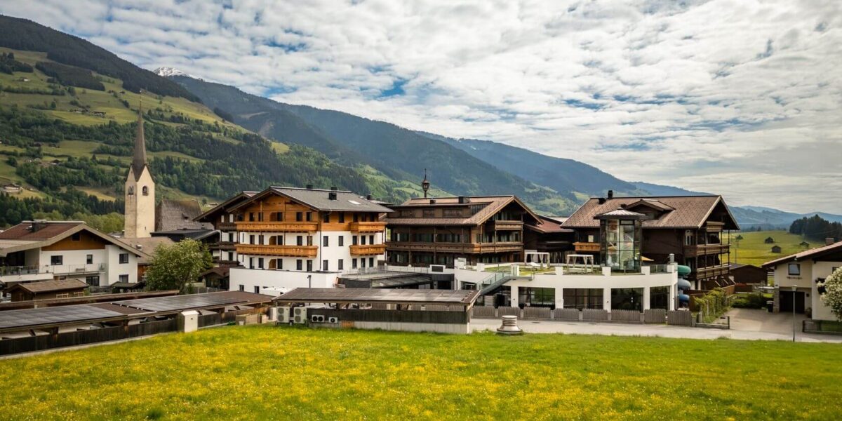Eine Ansammlung traditioneller alpiner Gebäude und eine Kirche mit einem hohen Kirchturm stehen neben grünen Feldern und Bergen unter einem teilweise bewölkten Himmel.