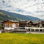 Eine Ansammlung traditioneller alpiner Gebäude und eine Kirche mit einem hohen Kirchturm stehen neben grünen Feldern und Bergen unter einem teilweise bewölkten Himmel.