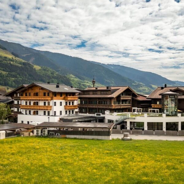Eine Ansammlung traditioneller alpiner Gebäude und eine Kirche mit einem hohen Kirchturm stehen neben grünen Feldern und Bergen unter einem teilweise bewölkten Himmel.