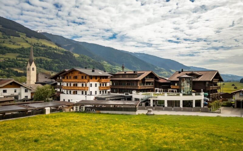 Eine Ansammlung traditioneller alpiner Gebäude und eine Kirche mit einem hohen Kirchturm stehen neben grünen Feldern und Bergen unter einem teilweise bewölkten Himmel.
