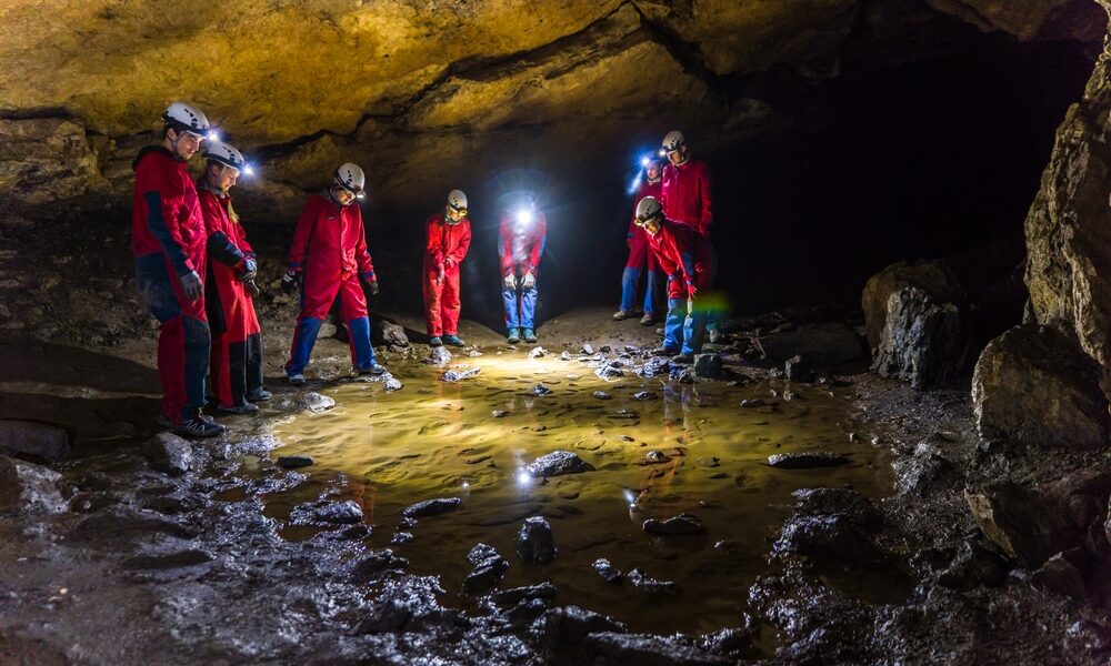 Eine Gruppe von Menschen in roten Anzügen und Helmen mit Scheinwerfern steht in einer Höhle um ein flaches Wasserbecken auf dem Höhlenboden herum.