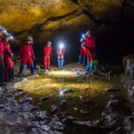 Eine Gruppe von Menschen in roten Anzügen und Helmen mit Scheinwerfern steht in einer Höhle um ein flaches Wasserbecken auf dem Höhlenboden herum.