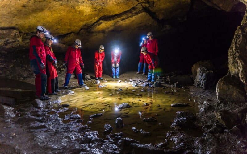 Eine Gruppe von Menschen in roten Anzügen und Helmen mit Scheinwerfern steht in einer Höhle um ein flaches Wasserbecken auf dem Höhlenboden herum.