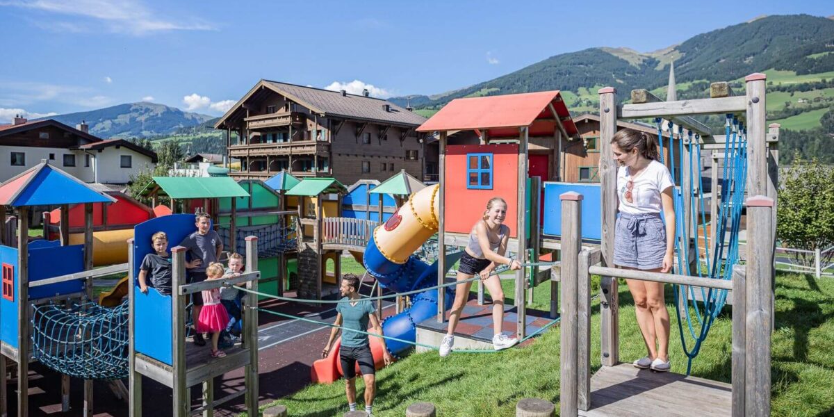 Kinder und ein Erwachsener spielen auf einem farbenfrohen Spielplatz im Freien, mit Gebäuden und grünen Hügeln im Hintergrund an einem sonnigen Tag.