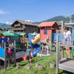 Kinder und ein Erwachsener spielen auf einem farbenfrohen Spielplatz im Freien, mit Gebäuden und grünen Hügeln im Hintergrund an einem sonnigen Tag.