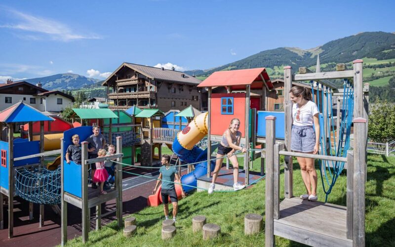 Kinder und ein Erwachsener spielen auf einem farbenfrohen Spielplatz im Freien, mit Gebäuden und grünen Hügeln im Hintergrund an einem sonnigen Tag.