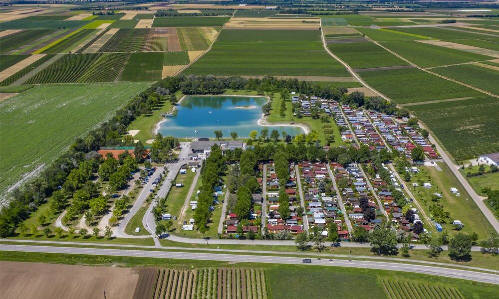 Luftaufnahme eines Campingplatzes mit Reihen von Wohnwagen und Hütten neben einem großen Teich, umgeben von Ackerland und Feldern unter einem klaren Himmel.