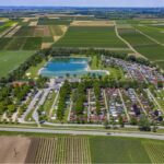 Luftaufnahme eines Campingplatzes mit Reihen von Wohnwagen und Hütten neben einem großen Teich, umgeben von Ackerland und Feldern unter einem klaren Himmel.