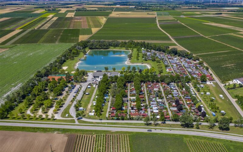 Luftaufnahme eines Campingplatzes mit Reihen von Wohnwagen und Hütten neben einem großen Teich, umgeben von Ackerland und Feldern unter einem klaren Himmel.