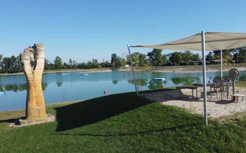Eine Parkszene mit einer großen Handskulptur an einem See, einem schattigen Sandkasten mit Spielgeräten und Grasflächen unter einem klaren blauen Himmel.