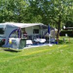 Ein Mann steht unter der Markise eines geparkten Wohnmobils auf einer Wiese mit Tischen, Stühlen, Wäschetrocknern und schattenspendenden Bäumen.