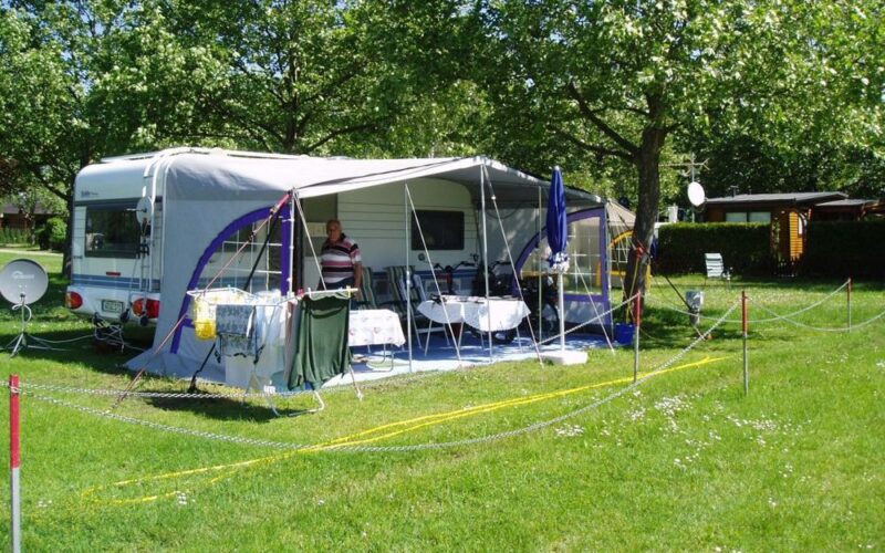 Ein Mann steht unter der Markise eines geparkten Wohnmobils auf einer Wiese mit Tischen, Stühlen, Wäschetrocknern und schattenspendenden Bäumen.