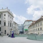 Ein geräumiger Innenhof mit historischen, blassen Gebäuden, Glasgeländern, Sitzgelegenheiten im Freien und einem modernen Gebäude im Hintergrund unter einem teilweise bewölkten Himmel.