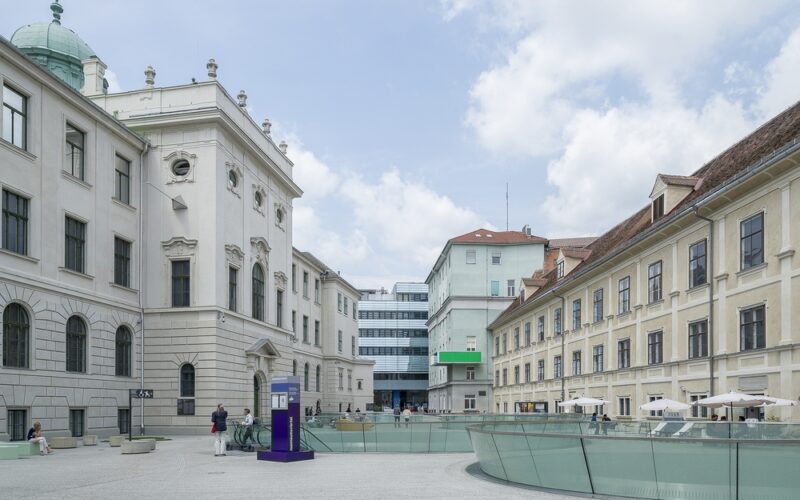 Ein geräumiger Innenhof mit historischen, blassen Gebäuden, Glasgeländern, Sitzgelegenheiten im Freien und einem modernen Gebäude im Hintergrund unter einem teilweise bewölkten Himmel.