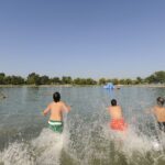 Vier Kinder rennen in einen See und planschen im Wasser, während im Hintergrund Bäume und ein klarer Himmel zu sehen sind.