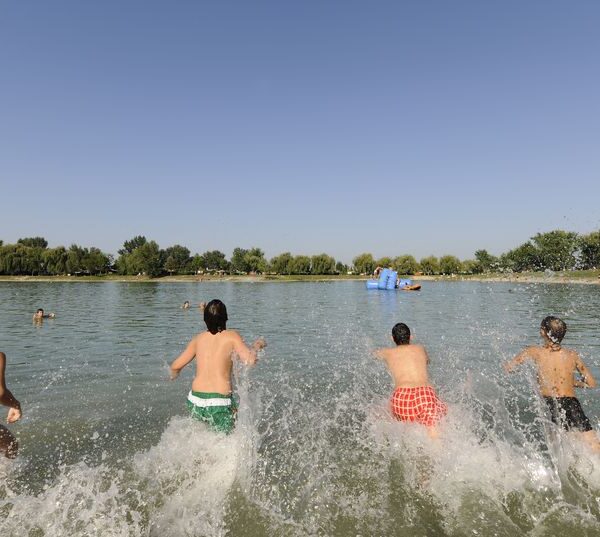 Vier Kinder rennen in einen See und planschen im Wasser, während im Hintergrund Bäume und ein klarer Himmel zu sehen sind.