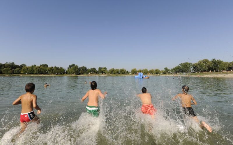 Vier Kinder rennen in einen See und planschen im Wasser, während im Hintergrund Bäume und ein klarer Himmel zu sehen sind.