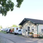 An einer Straße steht ein kleines Gebäude mit der Aufschrift "Camping-Büro", in dem mehrere Wohnmobile und Wohnwagen auf einem schattigen Stellplatz geparkt sind.