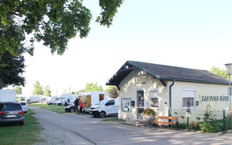 An einer Straße steht ein kleines Gebäude mit der Aufschrift "Camping-Büro", in dem mehrere Wohnmobile und Wohnwagen auf einem schattigen Stellplatz geparkt sind.