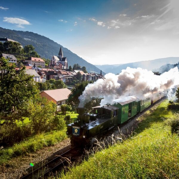 Ein Dampfzug fährt durch ein grünes Bergdorf mit Rauchschwaden, Häusern und einer Kirche im Hintergrund und bewaldeten Bergen in der Ferne unter einem teilweise bewölkten Himmel.