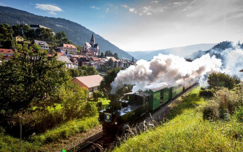 Ein Dampfzug fährt durch ein grünes Bergdorf mit Rauchschwaden, Häusern und einer Kirche im Hintergrund und bewaldeten Bergen in der Ferne unter einem teilweise bewölkten Himmel.