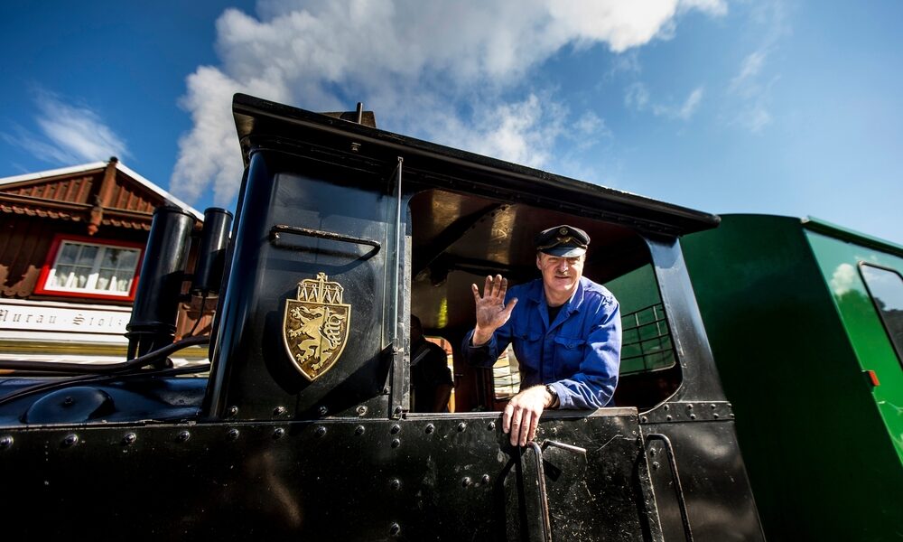 Ein Zugführer in blauer Uniform winkt aus dem Fenster einer schwarzen Lokomotive unter einem teilweise bewölkten Himmel.