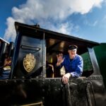 Ein Zugführer in blauer Uniform winkt aus dem Fenster einer schwarzen Lokomotive unter einem teilweise bewölkten Himmel.
