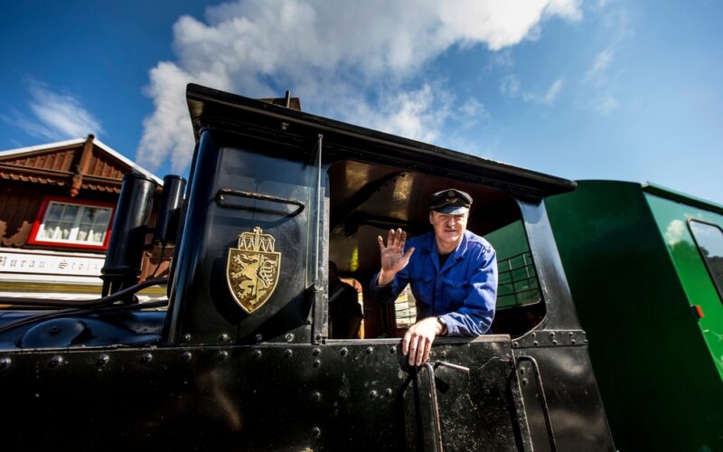 Ein Zugführer in blauer Uniform winkt aus dem Fenster einer schwarzen Lokomotive unter einem teilweise bewölkten Himmel.