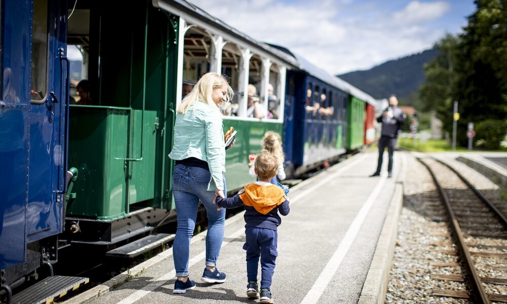 Eine Frau und zwei Kinder gehen an einem sonnigen Tag an einem Bahnsteig neben einem stehenden Personenzug entlang.