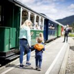 Eine Frau und zwei Kinder gehen an einem sonnigen Tag an einem Bahnsteig neben einem stehenden Personenzug entlang.