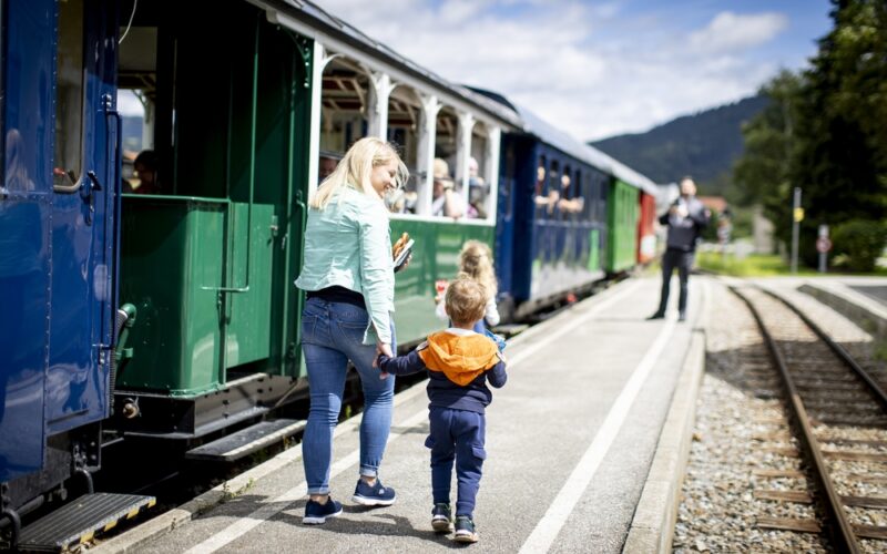 Eine Frau und zwei Kinder gehen an einem sonnigen Tag an einem Bahnsteig neben einem stehenden Personenzug entlang.