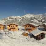Mehrere Holzhütten stehen in einer verschneiten Landschaft mit Bergen im Hintergrund unter einem klaren blauen Himmel.