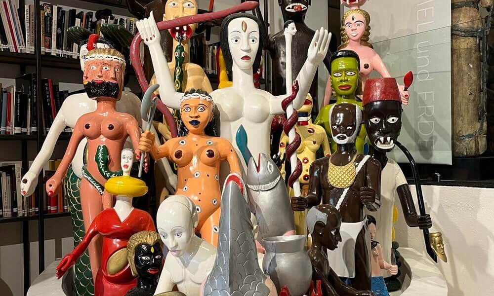 Eine Gruppe farbenfroher, stilisierter humanoider und tierischer Skulpturen ist in einer weißen, schalenartigen Struktur vor Bücherregalen ausgestellt.