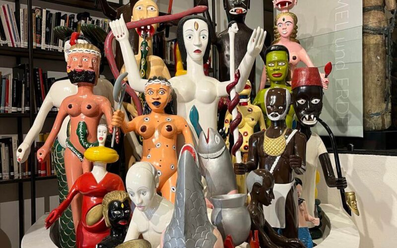 Eine Gruppe farbenfroher, stilisierter humanoider und tierischer Skulpturen ist in einer weißen, schalenartigen Struktur vor Bücherregalen ausgestellt.