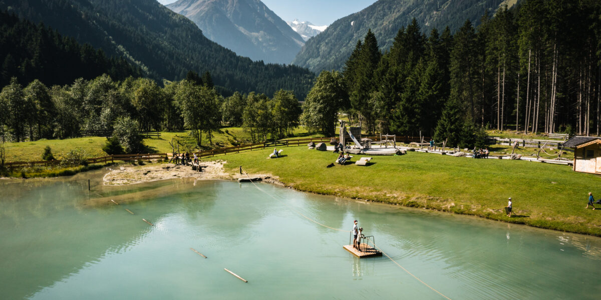 Menschen stehen auf einem kleinen Holzfloß auf einem ruhigen See, umgeben von Gras, Bäumen und Bergen unter einem strahlend blauen Himmel.