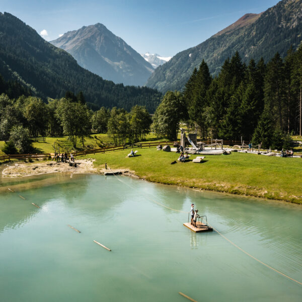Menschen stehen auf einem kleinen Holzfloß auf einem ruhigen See, umgeben von Gras, Bäumen und Bergen unter einem strahlend blauen Himmel.